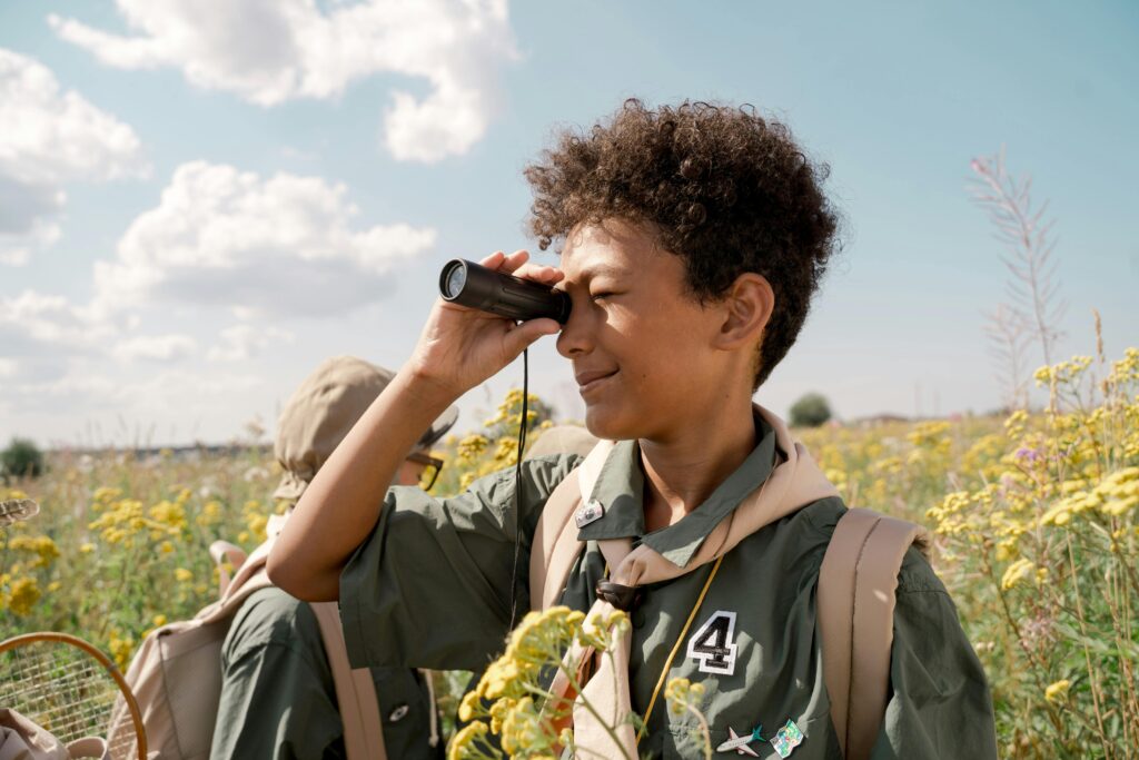 A boy scout observes the landscape through binoculars during a summer camp in a blooming field.