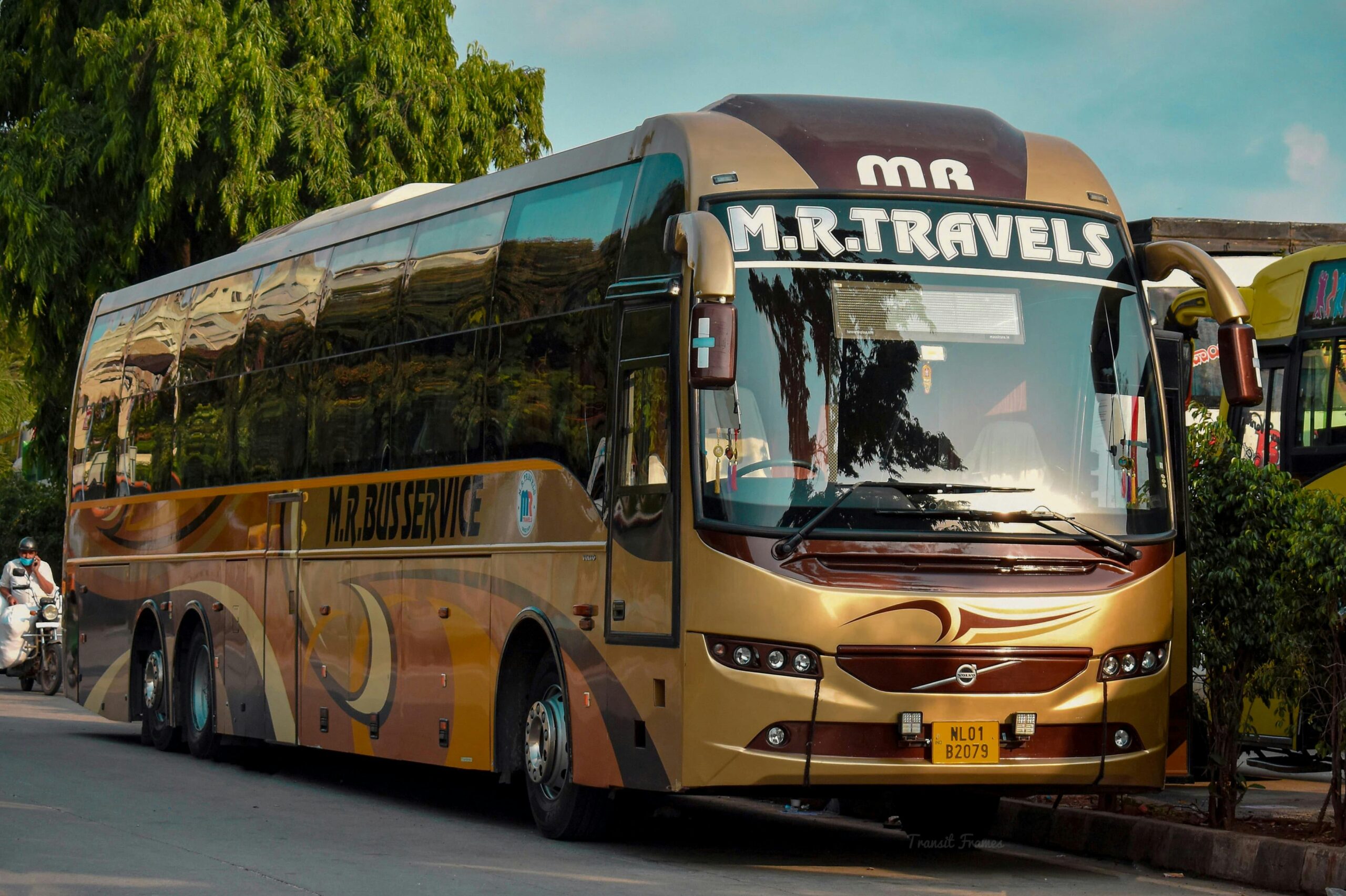 Golden luxury tour bus from M.R. Travels parked on street during the day with bright surroundings.